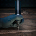 Schwarze und silberne ACUS M1 Grip Plus Tattoo-Maschine mit metallischer Nadel-TIP, gehalten in einer behandschuhten Hand für präzises Tätowieren