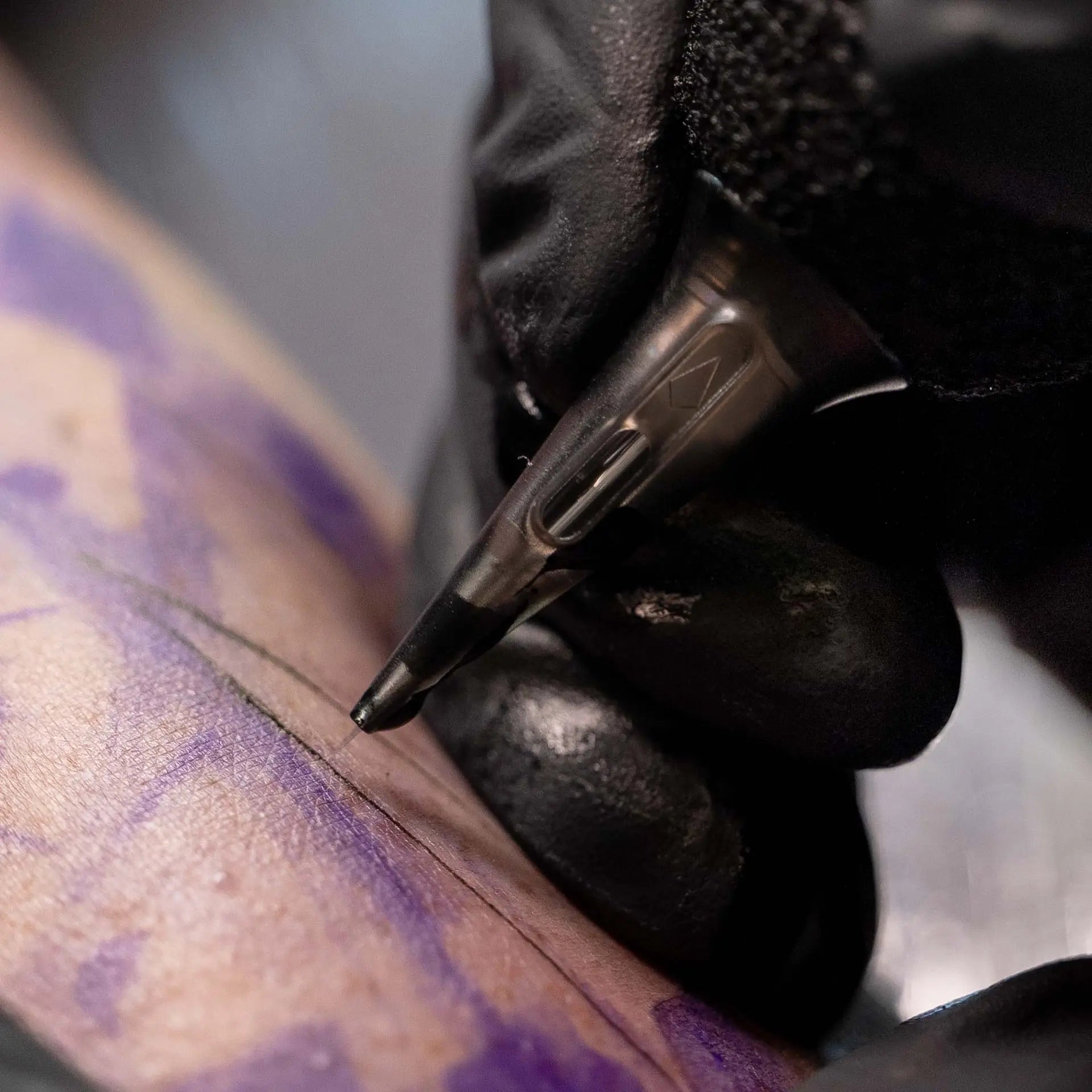Black piranha cartridge fine line premium tattoo needle held in gloved hand poised over purple ink skin outline