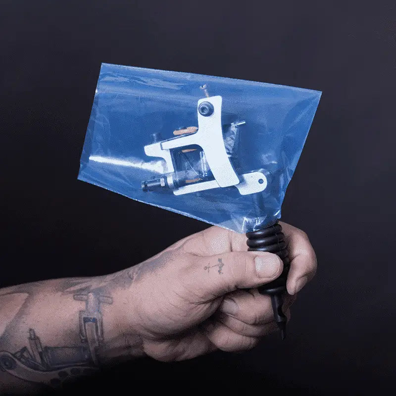 White tattoo machine encased in clear blue plastic bag held by tattooed hand for piranha machine bags product