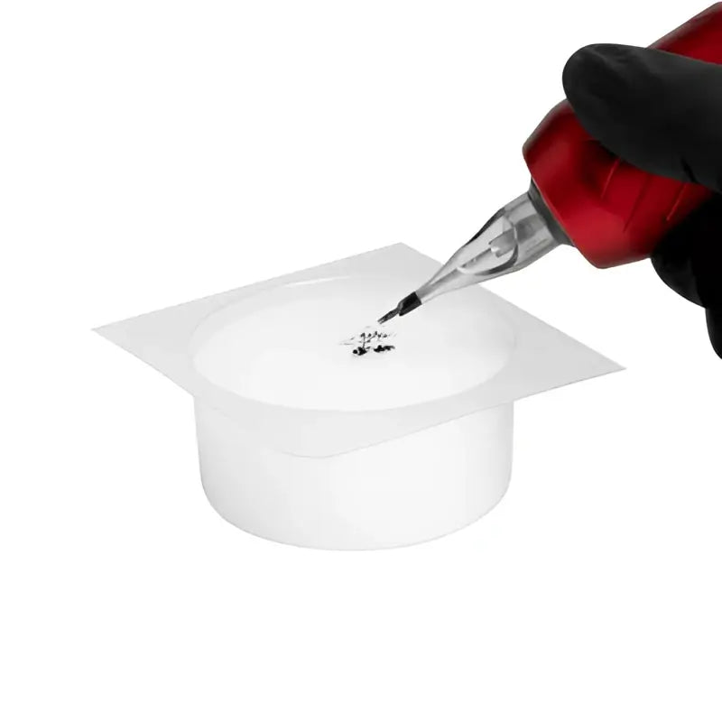 Rote Tätowiermaschine mit schwarzer Nadel tip trägt Farben auf weiße runde Spülkappe mit Schaum für saferly 24pcs Kit