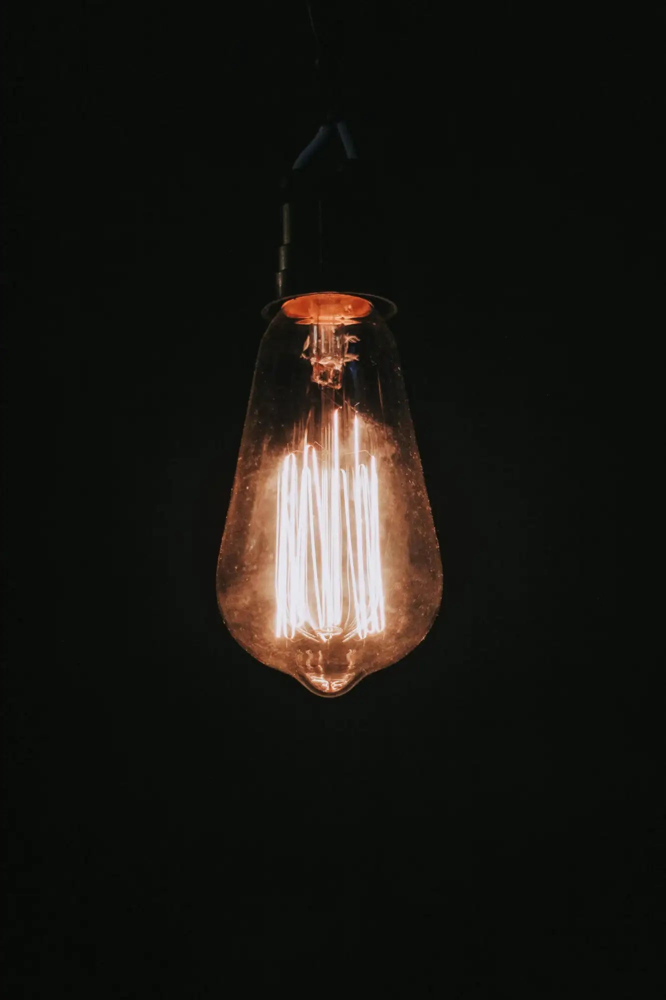 A glowing vintage-style incandescent bulb with a clear glass envelope and visible filament, suspended against a dark background.